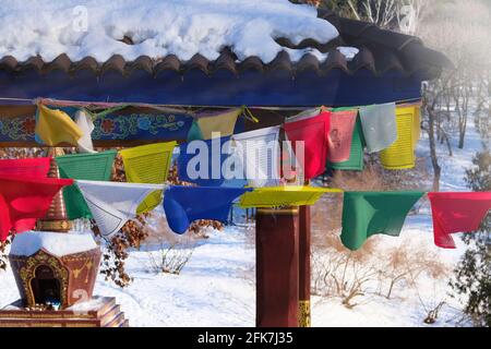 Buddhistische tibetische Gebetsfahnen fliegen im Winter auf dem Hintergrund von Bäumen auf Wind. Sonnenlicht sichtbar durch zahlreiche bunte Gebetsfahnen. Stockfoto