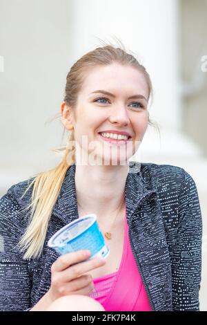 Schöne junge blonde Haare Mädchen lächeln und essen Joghurt in der Stadt, vertikal Stockfoto