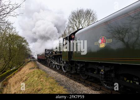 Historische Dampflok und Waggons auf Gleisen, Rauchwolken auf der historischen Eisenbahn (Mann auf der Fußplatte) - KWVR, Yorkshire, England. Stockfoto