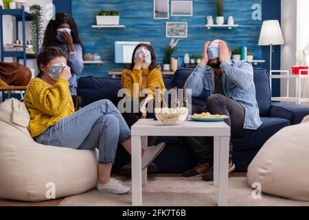 Eine Gruppe von multirassischen Freunden, die eine Horrorsendung im fernsehen anschauen, genießt es, gemeinsam eine Gesichtsmaske zu tragen, um eine Infektion mit Covid 19 während der globalen Pandemie zu verhindern und Spaß auf der Couch zu haben Stockfoto