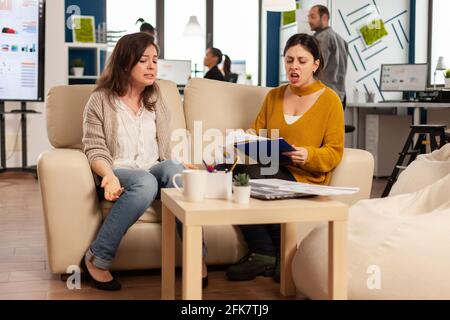 Eine Führungspersönlichkeit brüllt die Angestellte an, die in einem neuen Start-up-Geschäft auf der Couch sitzt, verärgert über eine schlechte Vertragsvereinbarung. Frauen, die große Konflikte haben, streiten über Fehler aus Projektdokumenten Stockfoto