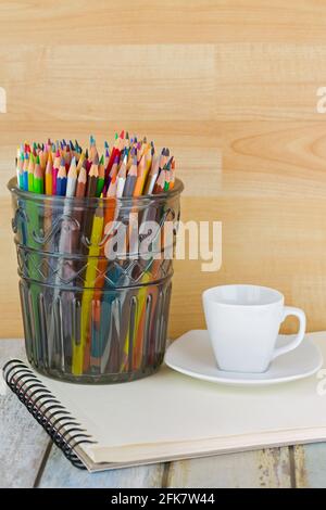 Heiße Espresso-Tasse neben großem Glas voller farbiger Bleistifte auf Skizzenzeichnung Buch. Stockfoto