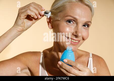 Nahaufnahme des Porträts einer reizenden, reifen blonden Frau, die lächelnd auf die Kamera lächelt, während sie saures Serum mit Pipette auf ihr Gesicht aufbringt und isoliert auf Beige posiert Stockfoto