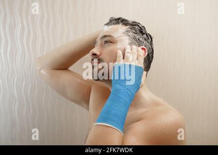 Gebrochenes Handgelenk in moderner blauer wasserdichter Bandage. Ein gutaussehender Mann mittleren Alters duscht mit in einen Gipsguss gewickelter Hand, wäscht seine Haare im Badezimmer Stockfoto