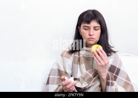 Porträt einer kranken attraktiven Frau, umhüllt von einer Decke, die einen Apfel, einen Kopierraum hält. Das Mädchen befindet sich in Quarantäne. Gesunde natürliche Ernährung Konzept Stockfoto