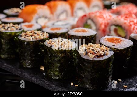 Nahaufnahme einer aufgeschnittenen Sushi-Rolle, die oben mit Sesamsamen auf einem schwarzen Brett auf dem Hintergrund anderer Sushi-Rollen bestreut wurde. Japanische orientalische Küche. Stockfoto