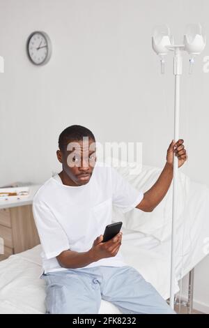 Vertikales Porträt eines jungen afroamerikanischen Mannes mit Smartphone im Krankenhauszimmer mit iv-Tropf-Setup, Kopierraum Stockfoto