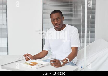 Porträt eines afroamerikanischen Mannes, der im Krankenhaus zu Mittag isst und die Kamera anschaut und den Raum kopiert Stockfoto