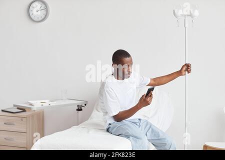 Porträt eines jungen afroamerikanischen Mannes mit Smartphone im Krankenhauszimmer mit iv-Tropf-Setup, Kopierraum Stockfoto