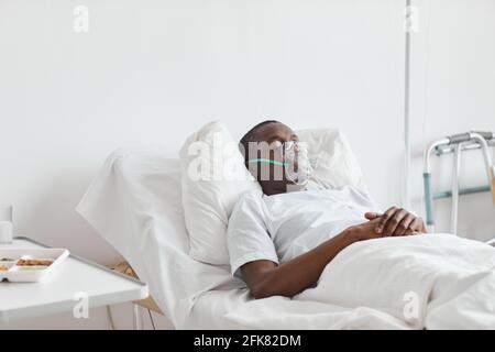 Porträt eines afroamerikanischen Mannes, der mit Sauerstoffmaske im weißen Zimmer auf einem Krankenhausbett schläft, Kopierraum Stockfoto