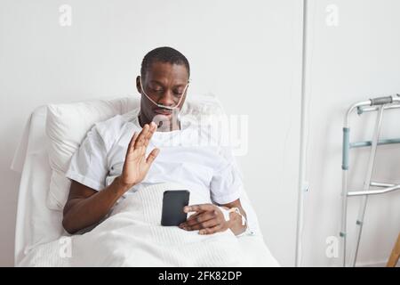 Porträt eines afroamerikanischen Mannes, der im Krankenhaus per Videoanruf spricht und zur Kamera winkt, während er im weißen Bett liegt und den Raum kopiert Stockfoto
