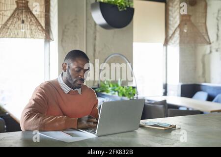 Porträt eines erwachsenen afroamerikanischen Mannes, der seinen Laptop benutzt, während er in einem modernen Café arbeitete, das mit frischen grünen Pflanzen und einem Kopierraum dekoriert ist Stockfoto