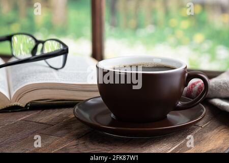 Tasse Kaffee und Buch auf einem rustikalen Holztisch An einem Fenster mit ländlichem Hintergrund Stockfoto