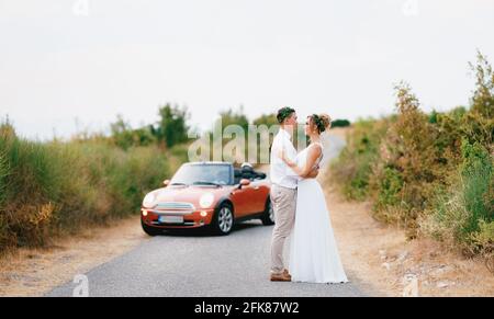 Die Braut und der Bräutigam stehen und umarmen sich in der Nähe der Auto auf der Autobahn in den Bergen und sieht zärtlich aus Aufeinander zu Stockfoto