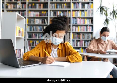 Multiethnische Studenten studieren während der Pandemie, sitzen an einem Tisch in der Ferne in einer schützenden medizinischen Maske in der Bibliothek mit einem Laptop, hören den Vortrag, afroamerikanisches Mädchen macht Notizen Stockfoto