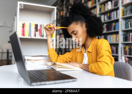 Smart nachdenklich konzentrierte gemischte Rennen Student Mädchen, sitzt am Tisch in der Universitätsbibliothek, über Projekt oder Hausaufgaben, Wissen zu gewinnen, Vorbereitung auf Prüfungen, Buch zu lesen Stockfoto