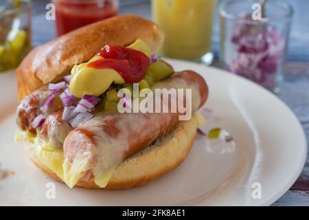 Nahaufnahme eines köstlichen und leckeren hausgemachten Hot Dog Burger mit Käse, Gurken, Zwiebeln, Senf und Ketchup auf einem Teller auf dem Küchentisch serviert Stockfoto