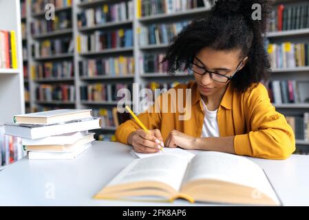 Erfolgreiche, intelligente junge Frau, afroamerikanische Studentin mit Brille, sitzt am Tisch in der Universitätsbibliothek, bereitet sich auf eine Vorlesung oder Prüfung vor, macht Hausaufgaben, schreibt Notizen in einem Notizbuch Stockfoto