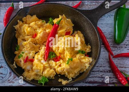 Würzige Rühreier mit Chilischoten, serviert in einer gusseisernen Pfanne, isoliert auf Holzgrund. Tabellenansicht von oben Stockfoto