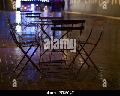 Ein verlassene Paar Tische mit Stühlen, draußen in einem Innenhof bei Winterregen, der den Boden nass gemacht hat und scharfe Neonreflexe schimmernd gesetzt hat. Stockfoto