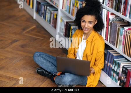 Nette, intelligente, fröhliche afroamerikanische Studentin mit lockigen Haaren in stilvoller Kleidung, die auf dem Boden in der Nähe des Bücherregals in der Bibliothek sitzt, einen Laptop hält, die Kamera anschaut und freundlich lächelt Stockfoto