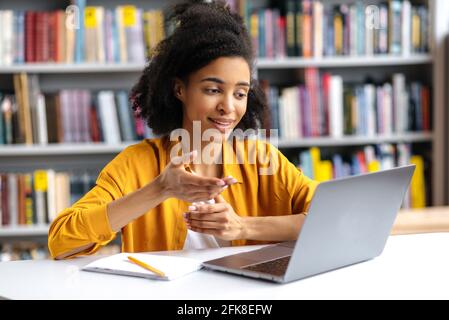 Online-Kommunikation. Selbstbewusste attraktive intelligente afroamerikanische Mädchen in stilvoller Kleidung, Studentin in der Universitätsbibliothek verwendet Laptop, reden auf Videokonferenz Freund oder Mitschülerin, gestikulierende Hand Stockfoto