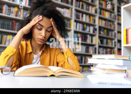 Smart afroamerikanische Studentin, sitzt in stilvoller Kleidung, in der Campus-Bibliothek am Tisch, Buch mit Interesse lesen, Vorbereitung auf Vorlesung oder Prüfung, Wissen und Lernkonzept Stockfoto