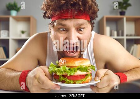 Hungriger, verrückter, molliger Mann in Sportkleidung, der Fast Food-Hamburger isst Hochformat Stockfoto