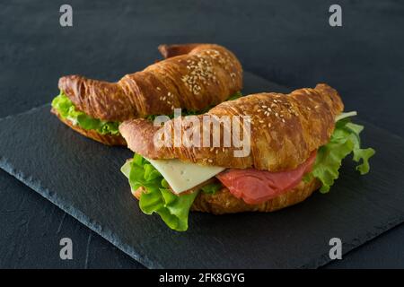 Croissant-Sandwiches mit gesalzenem Lachs auf einem Schreibtisch, serviert mit frischen Salatblättern, Rucola und Gemüse auf schwarzem Hintergrund. Stockfoto