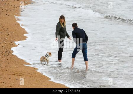 West Bay, Dorset, Großbritannien. April 2021. Wetter in Großbritannien. Ein Paar, das mit seinem Hund am Strand im Badeort West Bay in Dorset am Rande des Meeres paddelt, an einem Tag voller Wolken und warmer Sonneneinstrahlungen. Bildnachweis: Graham Hunt/Alamy Live News Stockfoto