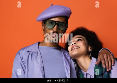 Trendy afroamerikanischen Mann in Baskenmütze und Sonnenbrille umarmt glücklich Frau auf Orange isoliert Stockfoto