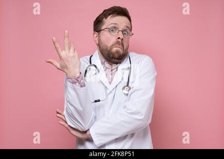 Der junge kaukasische Arzt macht eine Handbewegung in Verwirrung und sieht überrascht aus. Rosa Hintergrund. Stockfoto