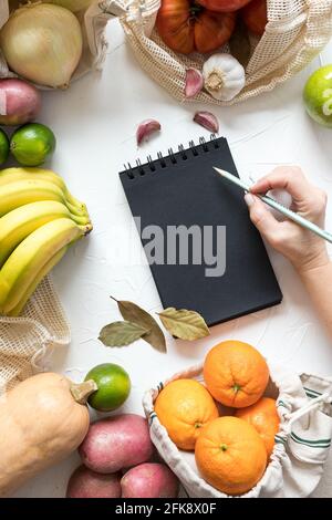 Frau schreibt eine Einkaufsliste auf einen schwarzen, spiralförmigen Notizblock, der von gesundem und rohem Gemüse umgeben ist. Stockfoto