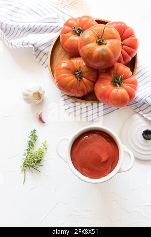 Blick von oben auf ein Holztablett voller frischer Tomaten auf einer rustikalen Serviette auf weißem Hintergrund. Daneben befindet sich ein weißer Auflauf mit Tomatensauce. Stockfoto