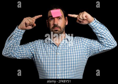 Bärtiger Mann in einem weißen und blauen karierten Hemd, der ein Papier hält, das an seiner Stirn klebte und mit den Fingern auf seinen Kopf zeigt, isoliert auf schwarzem Studio-Backgro Stockfoto