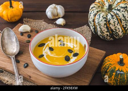 Schüssel mit Kürbissuppe auf einem Holzbrett auf dunkelbraunem rustikalem Holzhintergrund, verziert mit kleinen Kürbissen, Löffel und Samen Stockfoto