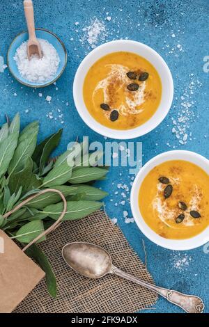 Zwei weiße Schüsseln mit Kürbis-/Karottensuppe, serviert mit Creme und Kürbiskernen auf blauem Hintergrund, verziert mit Salbei und Salz. Vertikale Ansicht von oben. Stockfoto