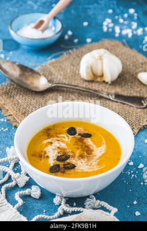 Kürbissuppe in einer weißen Schüssel mit Creme und Kürbiskernen auf blauem Hintergrund serviert. Vertikales Stockfoto. Stockfoto