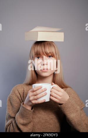 Ernsthaftes Gesichtskonzept. Nerd, Wissens- oder Bildungskonzept. Die attraktive kaukasische Universitätsstudentin trägt eine Brille und einen beigen Pullover mit Buch auf dem Kopf und hält eine Tasse Kaffee oder Tee in den Händen Stockfoto