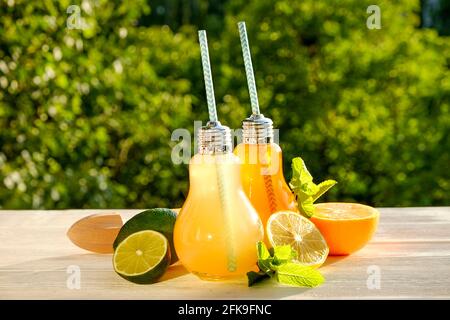 Zwei birnenförmige Gläser mit verschiedenen hausgemachten Limonaden, Scheiben aus Bio-Zitrone, Limette und Orange, Minze, Entsafter auf weißem Holztisch, ländliche Folia Stockfoto