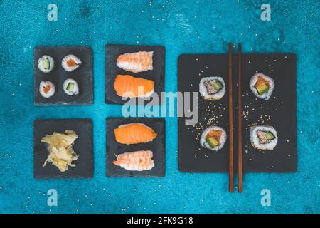 Verschiedene Nigiri- und Maki-Shushi-Sorten auf Schiefer, Ingwer und Holzstäbchen auf blauem Hintergrund, Draufsicht Stockfoto