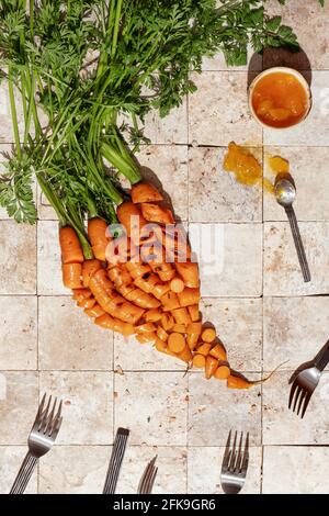 Ein Haufen gegrillter Karottenwurzeln mit grünen Blättern auf Stein Geschichten von Erdtönen mit Metallgabeln und Aprikosenmarmelade Draufsicht. Gesunde Ernährung und Diät fo Stockfoto