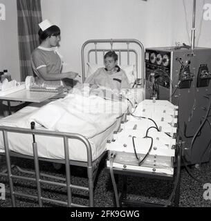 1960, historisch, eine Krankenschwester mit einem kleinen Reagenzglas, die neben einem jungen Jungen steht, der in einem Krankenhausbett mit einer Zeitung auf dem Schoß liegt. Er ist mit einer großen Maschine verbunden, einer Dialysegerät, die Blut durch einen Filter reinigt, der als Dialysator bekannt ist, South East London, England, Großbritannien. Dialyse ist eine medizinische Behandlung, die das Blut mit einem Gerät filtert und reinigt, was hilft, den Körper im Gleichgewicht zu halten, wenn die Nieren diese Funktion nicht erfüllen können. Stockfoto