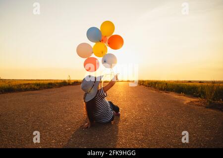 Kleines Mädchen sitzt auf der Straße und spielt mit bunten Ballons bei Sonnenuntergang. Rückansicht Stockfoto