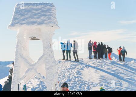 Kasprowy Wierch, Polen 28.01.2021 - EINE Gruppe von Bergsteigern auf einem verschneiten Berggipfel. Nahaufnahme Blick auf einen verschneiten Glockenturm. Landschaftlich klarer weißer Himmel im Hintergrund. Menschen, die Fotos von der Aussicht machen. Stockfoto