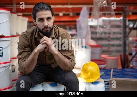 Porträt eines selbstbewussten, gut aussehenden jungen bärtigen Aktienkontrollers, der mit sitzt Umklammten die Hände im Lager der Lack- und Farbenfabrik Stockfoto
