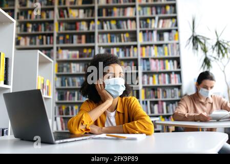 Zwei smarte Studentinnen verschiedener Nationalitäten in medizinischen Masken sitzen an Tischen in der Universitätsbibliothek, bereiten sich auf den Unterricht oder eine Prüfung vor, ein afroamerikanisches Mädchen schaut nachdenklich weg Stockfoto