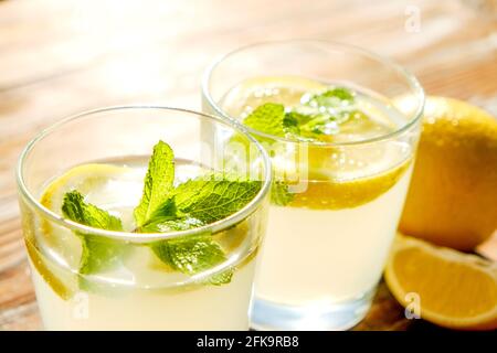 Zwei Gläser voll eiskaltem, erfrischendem Limonadengetränk mit Minzblättern im Sonnenlicht auf braunem, grunziertem Holztisch, Scheiben reifer Bio-Zitrone, wh Stockfoto