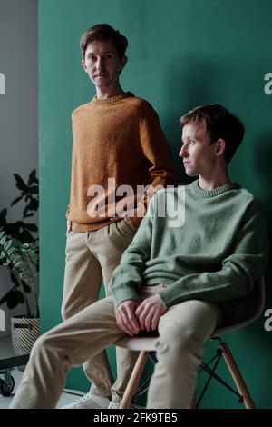Porträt eines hübschen jungen Mannes in einem Pullover, der die Hände hält In den Taschen und sein Zwillingsbruder sitzt auf Stuhl und Wegschauen Stockfoto