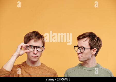 Junger blonde Mann in olivfarbenem Pullover, der Bruder beim Anpassen ansieht Brille vor orangefarbenem Hintergrund Stockfoto